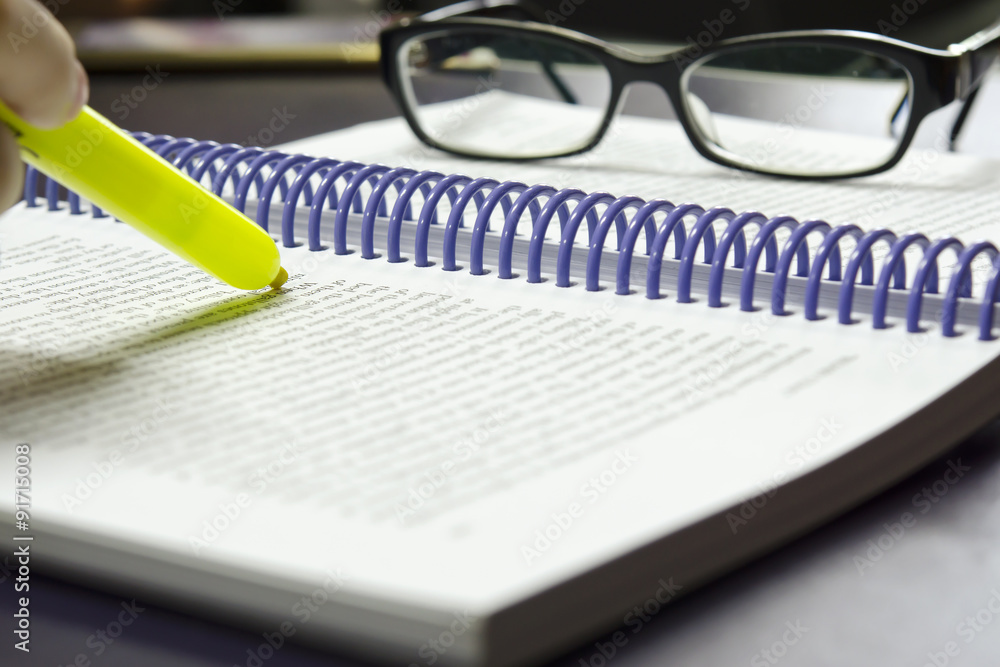 Hand holding highlighter pen highlights keywords on the book with eye glass in the background Hand holding highlighter pen highlights keywords on the book with eye glass in the background