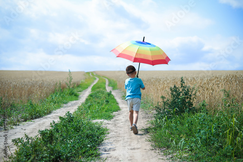 Little boy with big umbrella walking away on country road