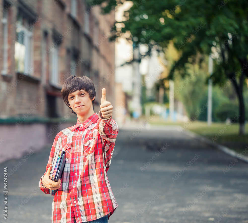 Fototapeta premium Smiling young student man holding a book, tablet and thumbs up against a city background