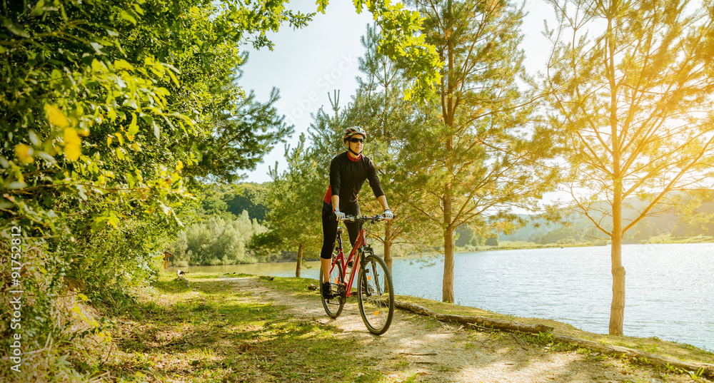 Obraz premium Young woman riding a bike in the forest next to a lake at sunset.