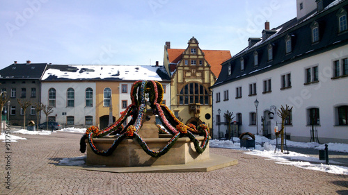 Osterbrunnen/Auf dem Marktplatz einer Kleinstadt in Thüringen ist zu Ostern der Brunnen mit vielen bunten Ostereiern geschmückt, im Hintergrund Häuser und das Rathaus.