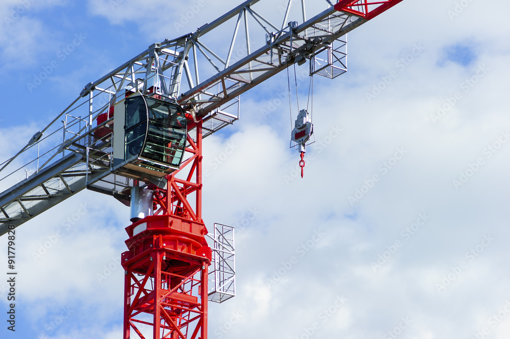 Red construction tower crane with grey jib and hook isolated on blue sky with white clouds background, detail 