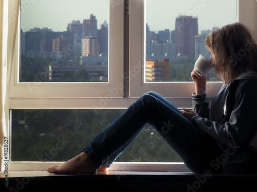 Девушка сидит на подоконнике и смотрит на город за окном. Пьет из чашки чай или кофе