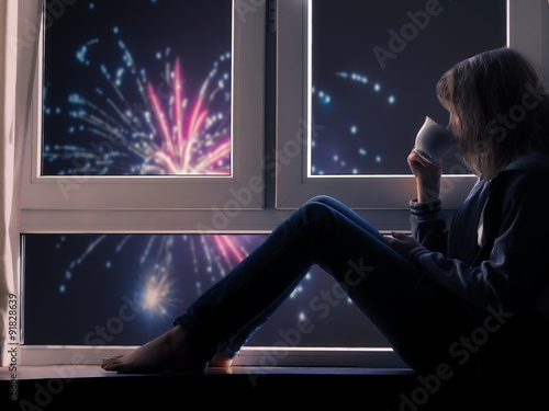 Девушка смотрит в окно на салют и пьет чай или кофе