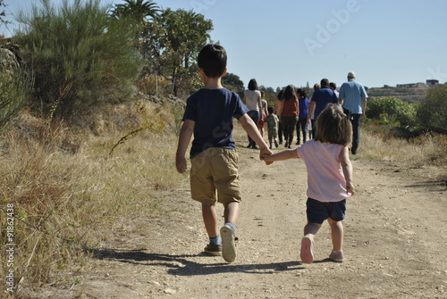 A boy and a girl holding hands, walking along a dirt road following a group of adults