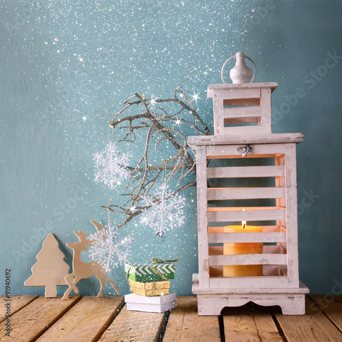 white wooden vintage lantern with burning candle, wooden deer, christmas gifts and tree branches on wooden table. retro filtered image with glitter overlay 