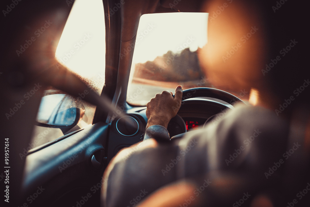 Naklejka premium Mężczyzna prowadzący samochód, ręka na kierownicy, patrząc na drogę przed zachodem słońca.