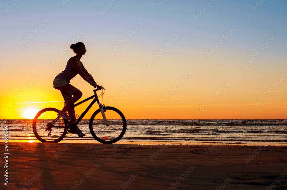 Fototapeta premium Silhouette of sporty woman riding bicycle on multicolored sunset