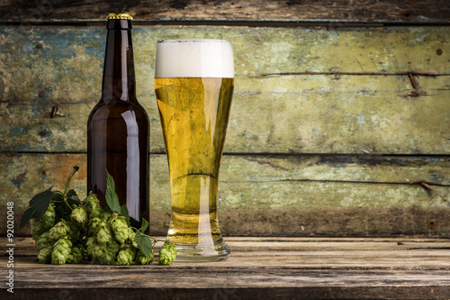 Bottle and mug full of beer with bunch of hops on wood background