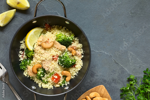 Couscous dish with chicken, broccoli, tomato, cashew nuts. Lemon, cashew nuts and parsley on the side, photographed overhead on slate with natural light. photo