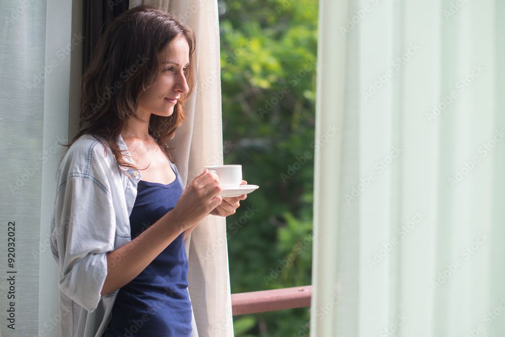 Woman in wide, long shirt is drinking tea or coffe and looking Woman in wide, long shirt is drinking tea or coffe and looking
