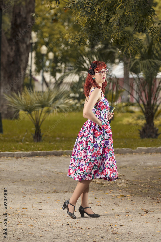 View of pinup young woman in vintage style clothing walking on the city park with a book.