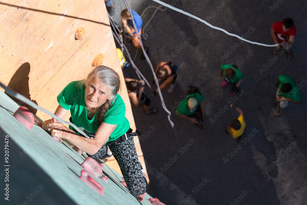 Fototapeta premium Aged Lady Doing Extreme Sport Elderly Female Moving Up on Outdoor Climbing Wall Sporty Clothing on Fitness Training Intense but Positive Face Using Rope and Belaying Gear