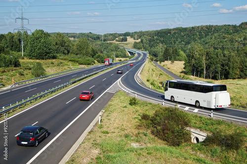 White Bus arriving to the asphalt highway on the slip road in a wooded landscape. Red passenger cars and truck driving on the highway. Electronic toll gate in the distance. View from above.