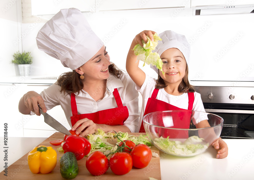 young mother and little daughter at house kitchen preparing salad for lunch wearing apron and cook hat young mother and little daughter at house kitchen preparing salad for lunch wearing apron and cook hat