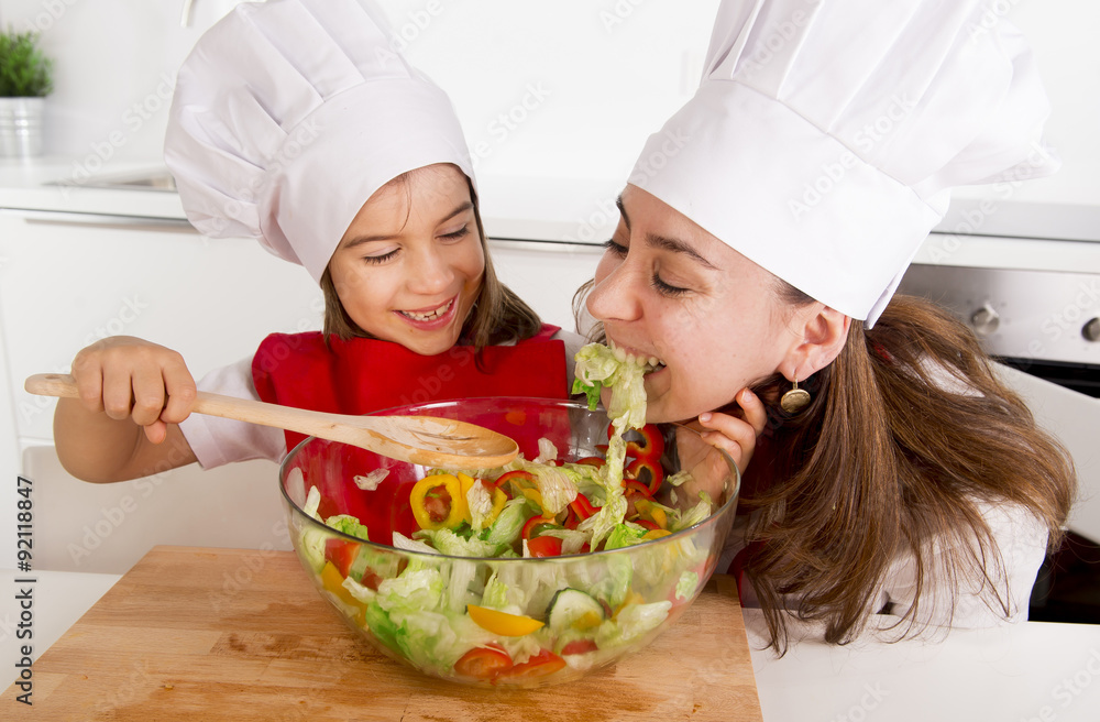 happy mother and little daughter at home kitchen preparing salad in apron and cook hat