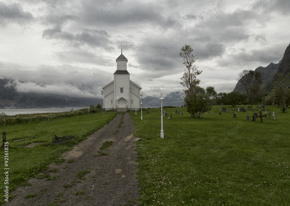 A walk way to the front of the church with some gravestones and light poles. Mountains and low haning clouds