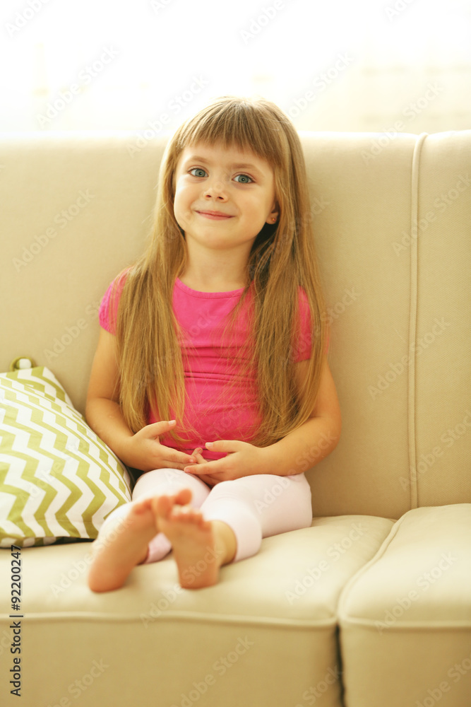 Beautiful little girl sitting on sofa, on home interior background Beautiful little girl sitting on sofa, on home interior background
