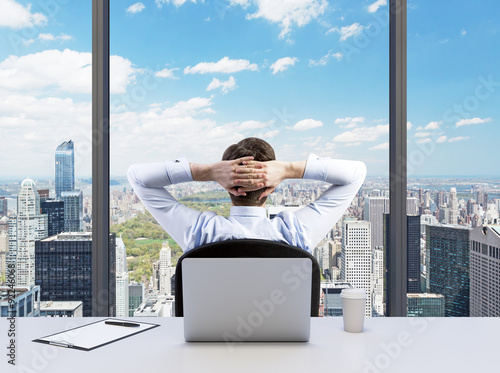Rear view of the relaxing businessman with crossed hands behind his head, who is looking at the Cntral park. Modern Panoramic office or work place with New York city view.