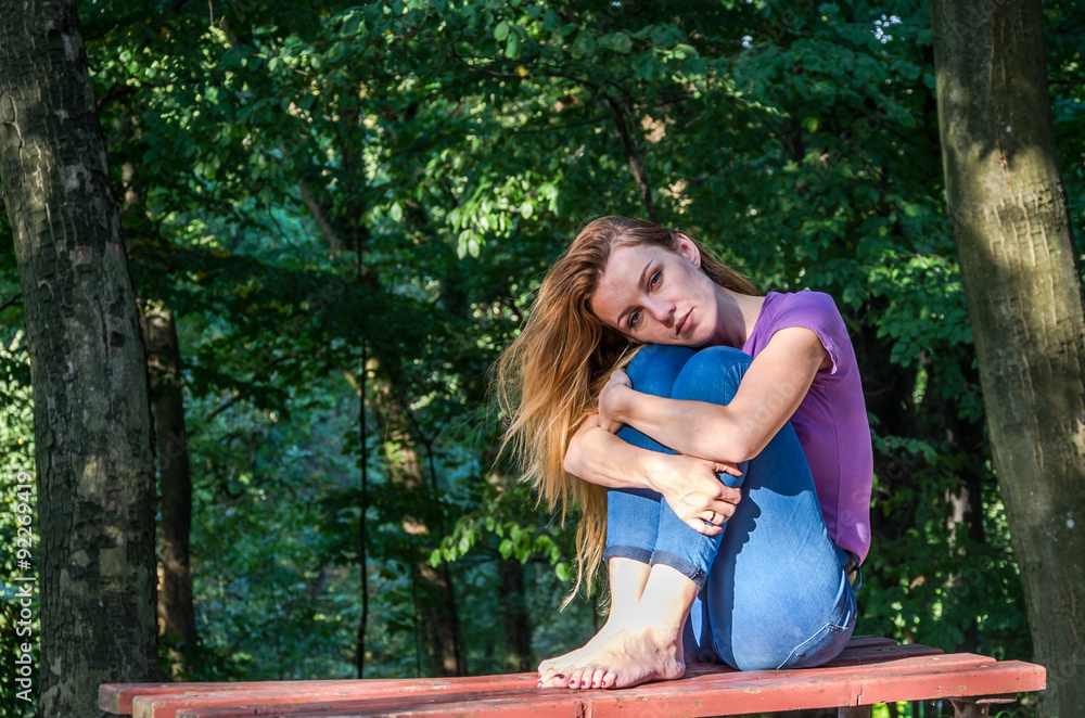 Young beautiful girl model in jeans and a T-shirt with long blond hair and sad smiles pensively posing for a walk in the autumn park sitting on a bench among the trees and vegetation Young beautiful girl model in jeans and a T-shirt with long blond hair and sad smiles pensively posing for a walk in the autumn park sitting on a bench among the trees and vegetation