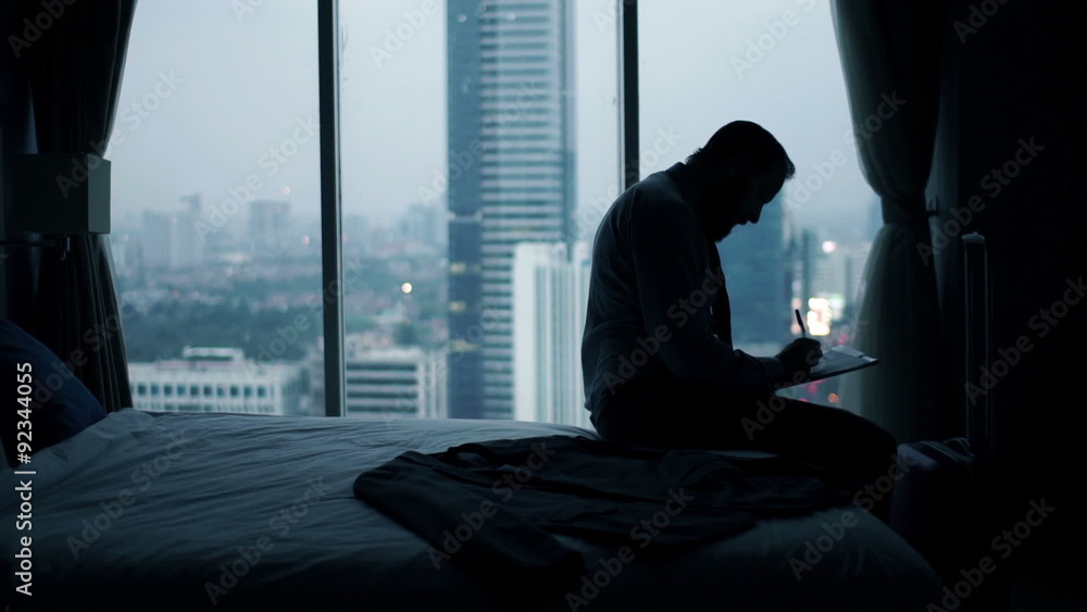 Young businessman writing notes in documents sitting on bed at home Young businessman writing notes in documents sitting on bed at home
