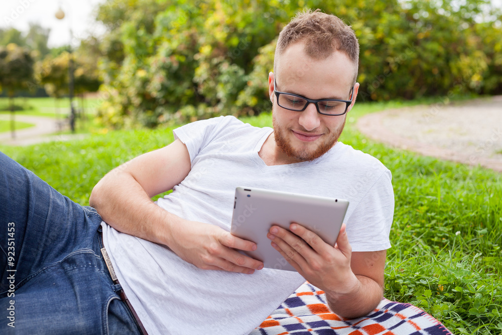 Bearded young man lying in park on blanket. He is using white tablet pc. Bearded young man lying in park on blanket. He is using white tablet pc.