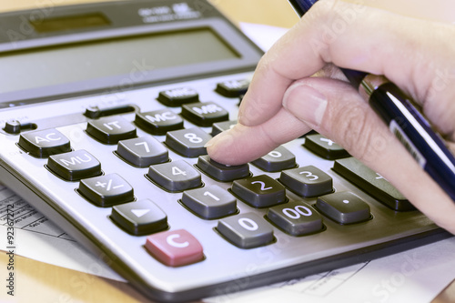 Photo of hand holding pen and pressing calculator buttons over documents photo