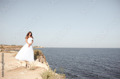 Young beautiful woman stading at seashore. Wearing white wedding dress. Bride posing outdoors. Wedding day. Celebration.