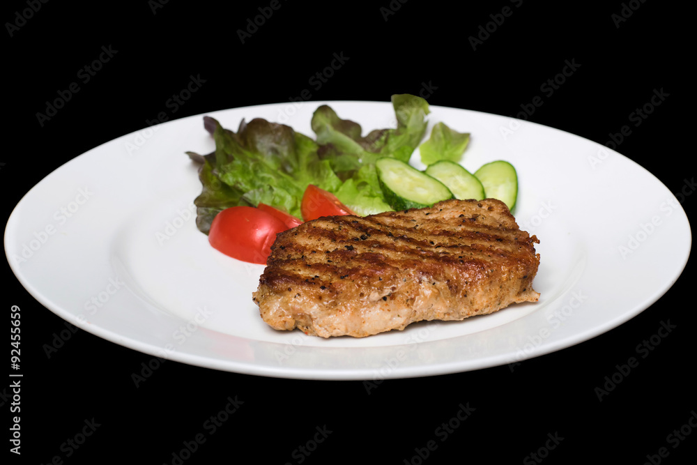 Photo of a pork stake on a white plate with a cucumber, tomato a