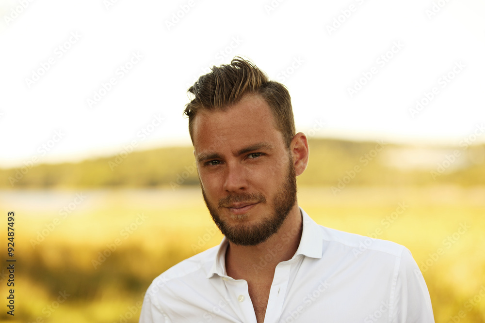 An attractive man in his 20s wearing a white shirt standing outside on a sunny summer day, with a big yellow field behind him. An attractive man in his 20s wearing a white shirt standing outside on a sunny summer day, with a big yellow field behind him.