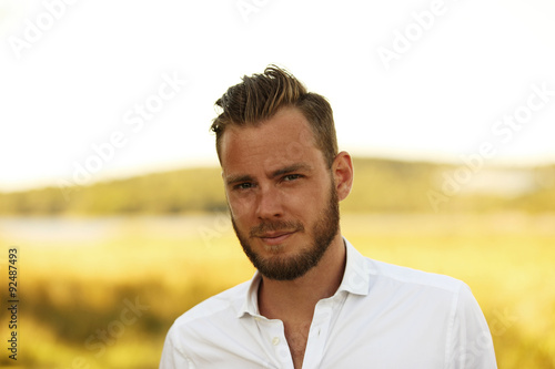 An attractive man in his 20s wearing a white shirt standing outside on a sunny summer day, with a big yellow field behind him.