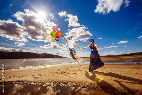 Happy young pair with bright balloons is jumping at summer beach