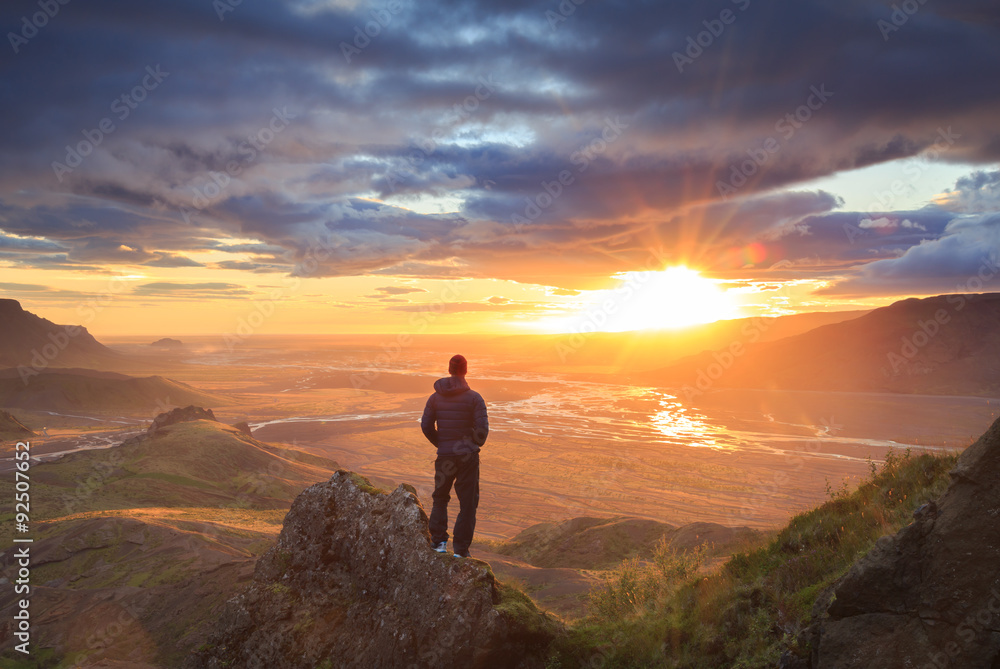 Hiker standing on a ledge of a mountain, enjoying the beautiful sunset over a wide river valley in Thorsmork, Iceland.
