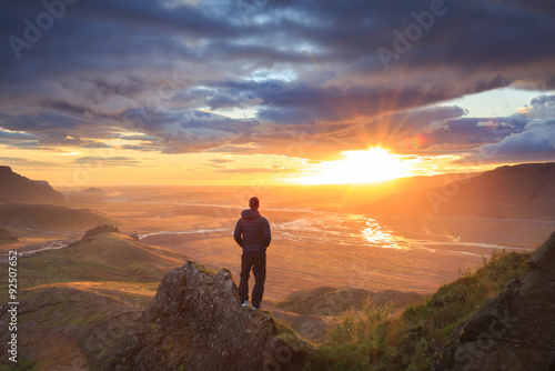 Hiker standing on a ledge of a mountain, enjoying the beautiful sunset over a wide river valley in Thorsmork, Iceland.
