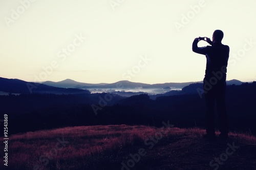 Hiker is taking photo of sunrise by smart phone on meadow. View to hilly landscape.