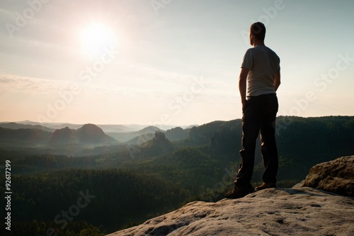 Satisfy tall hiker in grey shirt and dark trousers. Sprtsman on the peak of sharp rock edge watching down to landscape.