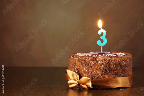 Birthday chocolate cake with burning candle as a number three on brown background