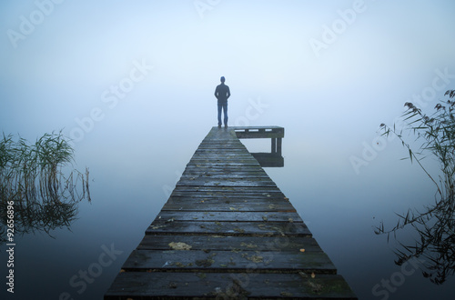 Canvas Print Man standing on a jetty at a lake during a foggy, gray morning.