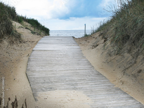 Sand dune landscape with wooden boardwalk leading towards sea and sky at Skrea Strand on a sunny day with dark clouds in Falkenberg, Sweden.