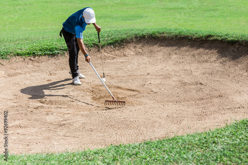 Yong asia golfer raking bunker after shot.