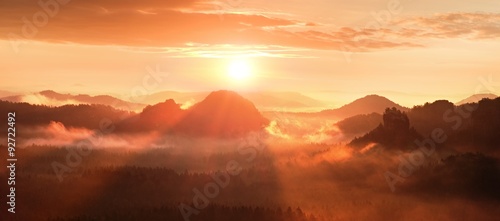 Red misty daybreak in a beautiful hills. Peaks of hills are sticking out from foggy background, red and orange fog below Sun rays.