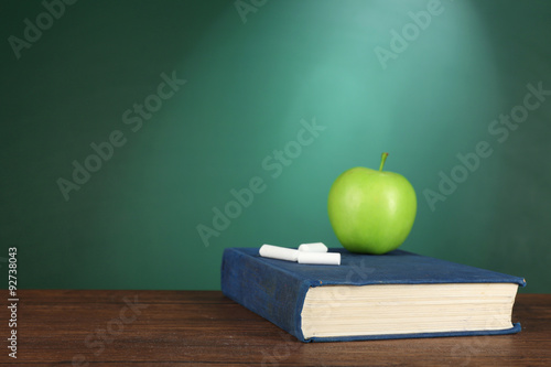 Book with green apple and pieces of chalk on green chalkboard background