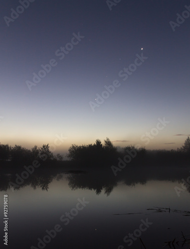 Riverside just before sunrise. Trees reflected in the water. Stars in the sky.