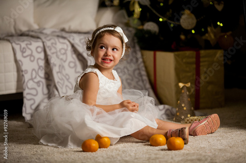 Cute happy little girl in beautiful white dress sitting on the carpet next to the christmas tree. Christmas tree with lights and gift boxes on the background. She is smiling and opening a gift box 