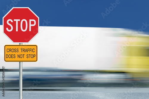Red stop road sign, motion blurred truck vehicle traffic in background, regulatory warning signage octagon, white octagonal frame, metallic pole post, yellow cross traffic does not stop text signage