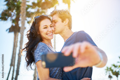 man kissing his girlfriend on the cheek for romantic selfie with lens flare and palm trees in background