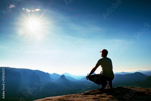 Runner in red cap and in dark sportswear. Man sit in squatting position on a rock in heather bushes, enjoy autumn scenery