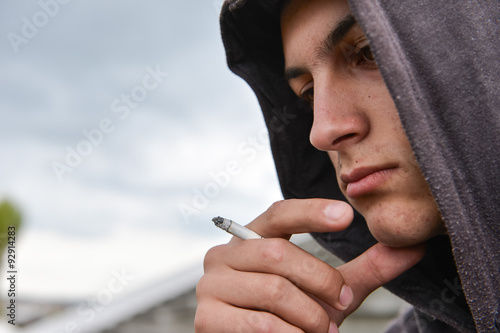 pensive and worried teenage boy with black hoodie is smoking cig