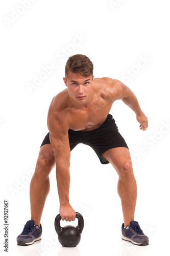 Fit man in sport shorts and sneakers picking up kettlebell from the floor. Full length studio shot isolated on white.