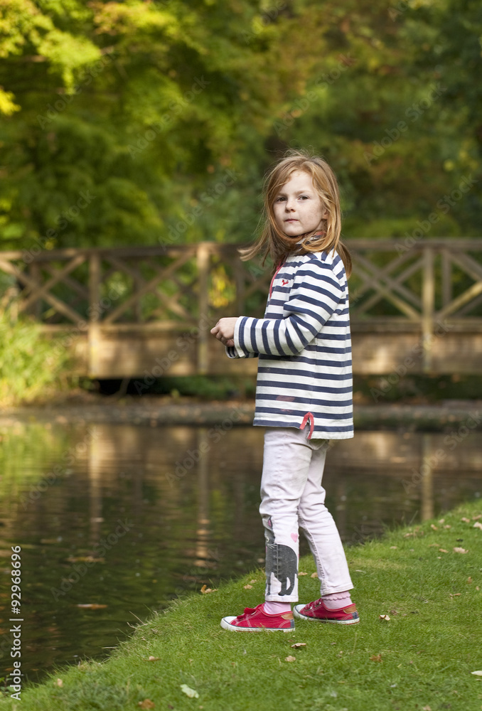 Fototapeta premium Young Girl Looking to Feed the Ducks. A young caucasian girl with ginger hair, is looking to feed the ducks on an ornamental pond in a park. There are vague signs of autumnal colour in the background.
